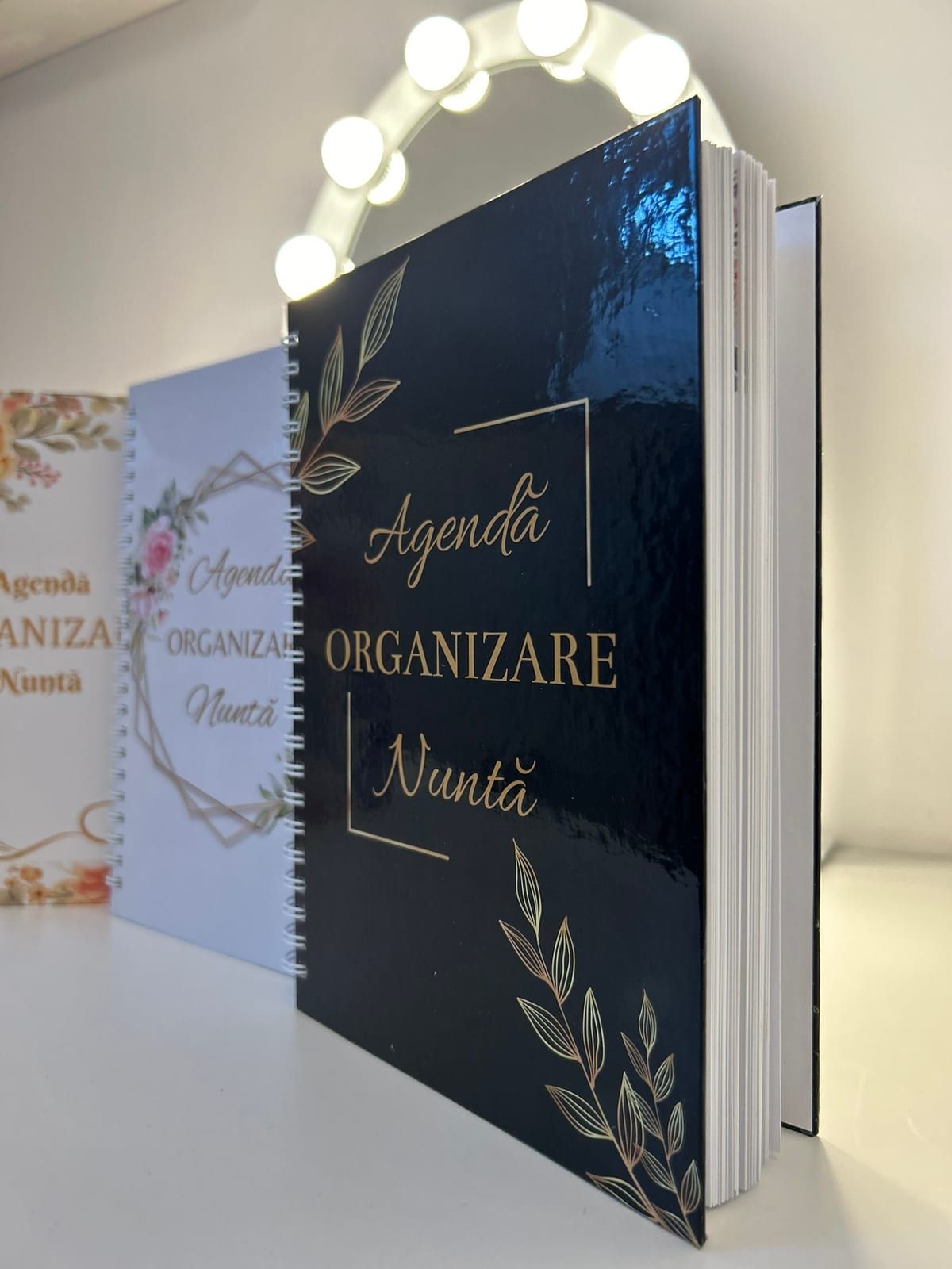 Agenda cu coperta neagră, deschisă lângă oglinda cu becuri